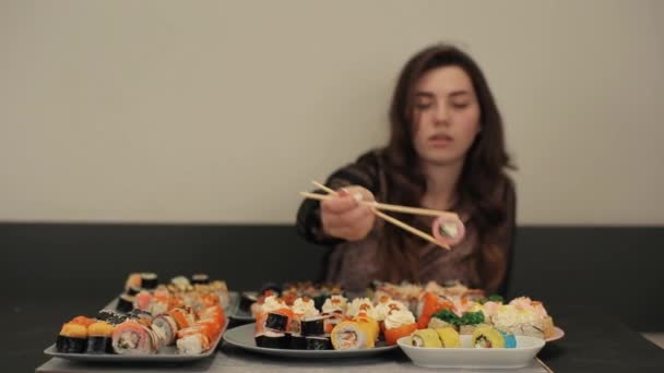 Una joven alegre se sienta en una mesa y mantiene los rollos de sushi con palillos a la altura de los ojos. Primer plano. La cámara cambia de enfoque — Vídeo de stock