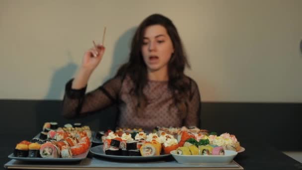 Энергичная молодая девушка сидит за столом в кафе, загроможденном тарелками суши, и выбирает, какой рулон ей съесть. Крупный план — стоковое видео