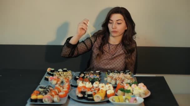 Nettes junges Mädchen sitzt an einem Tisch mit einer großen Auswahl an Sushi und lacht, als eines der Brötchen aus den Stäbchen fällt. Nahaufnahme — Stockvideo