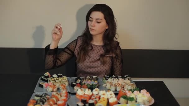 Ασιατική κουζίνα. Η νεαρή κοπέλα φέρνει στο στόμα της ένα ρολό επιλεγμένο από τα πολλά είδη σούσι που στέκονται στο τραπέζι του καφέ. Κοντινό πλάνο — Αρχείο Βίντεο