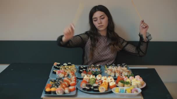 Молодая веселая девушка сидит за столом с большим количеством суши и веселыми палочками для еды. Имитация игры на барабане. Крупный план — стоковое видео