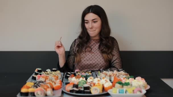 Una giovane donna caucasica felice mangia un rotolo e sorride con piacere. Ci sono un sacco di piatti di sushi diversi sul tavolo. Primo piano — Video Stock