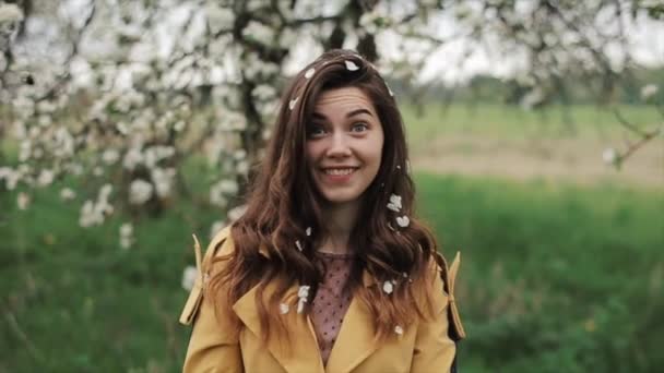 Una joven hermosa y emocional se levanta sobre el fondo de un árbol floreciente hace una cara sorprendida y sacude los pétalos del cabello. Primer plano. En cámara lenta. — Vídeos de Stock