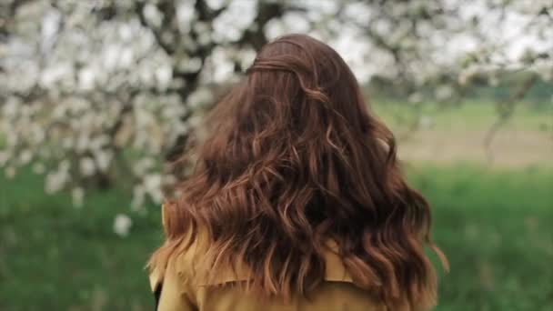 Een leuk jong meisje met lang haar draait naar de camera in de lentetuin met afbrokkelende bloemblaadjes. Een close-up. Langzame beweging — Stockvideo