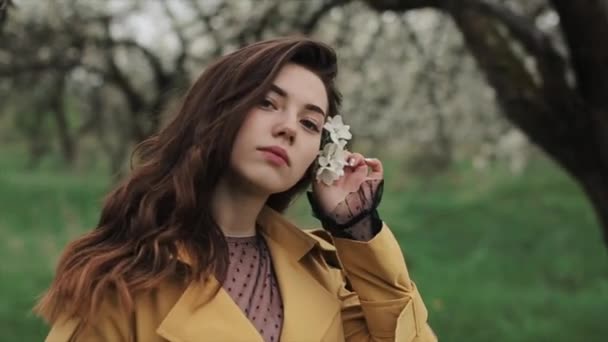 Κοντινό πλάνο ενός νεαρού ευγενικού κοριτσιού με ένα λουλούδι στα μαλλιά της στο φόντο των ανθισμένων δέντρων. Ανοιξιάτικη διάθεση Αργή κίνηση — Αρχείο Βίντεο