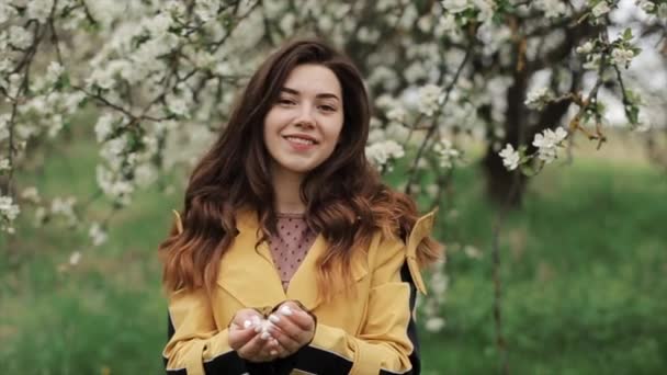 A nice smiling young girl is standing in the spring garden and blowing off the petals of white flowers from her palms. Close-up. Slow motion — Stock Video