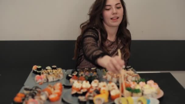 Молодая привлекательная девушка садится за стол с большим количеством тарелок с различными видами суши-роллов и наслаждается вкусом. Камера меняет фокус. Крупный план — стоковое видео