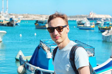 Teknelerin limanında su kenarında oturan genç adam, renkli Malta deniz manzaralı. Turist konsepti. Boşluğu kopyala.