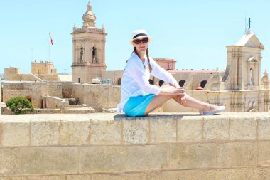 Young stylish woman in hat traveling in the Mdina city, Malta. Tourist concept. Copy Space. Blond woman resting. clipart
