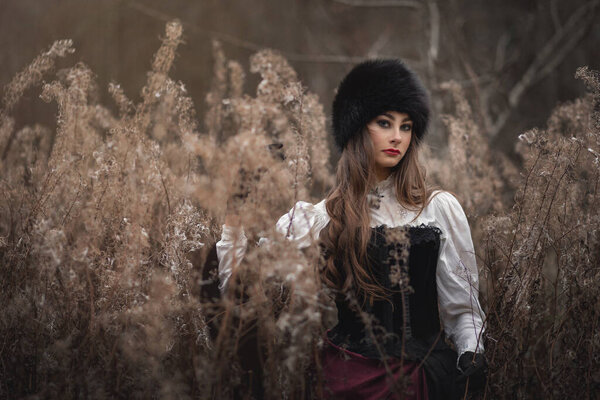 A mysterious young girl in a long dress with a train. The wizard leaves in a forest covered with fog. A background of trees with a haze away. Woman with retro costume and retro hat.