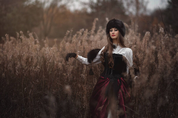 A mysterious young girl in a long dress with a train. The wizard leaves in a forest covered with fog. A background of trees with a haze away. Woman with retro costume and retro hat.