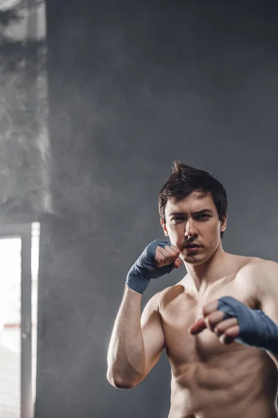 Strong boxer in the rays of the sun and smoke training hands with boxing wraps