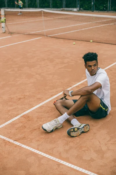 Young man in sports uniform sits on the ground of a tennis court