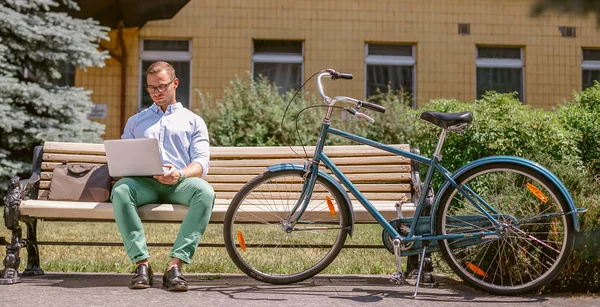 Бизнесмен, работающий на ноутбуке, сидит на скамейке, рядом с велосипедом — стоковое фото