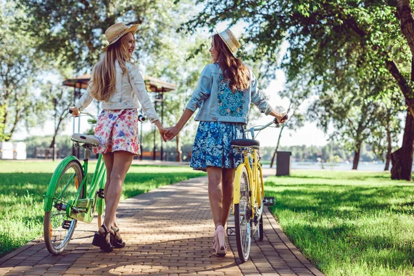 Rear view of two women holding hands walking in a park with bicycles.
