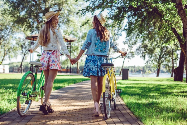 Rear view of two women holding hands walking in a park with bicycles.