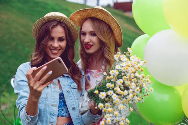 Jovens mulheres bonitas com balões e flores silvestres fazendo selfie no parque . — Fotografia de Stock