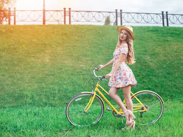 Retrato completo de una hermosa mujer rubia posando cerca de una bicicleta amarilla en un parque de césped verde — Foto de Stock