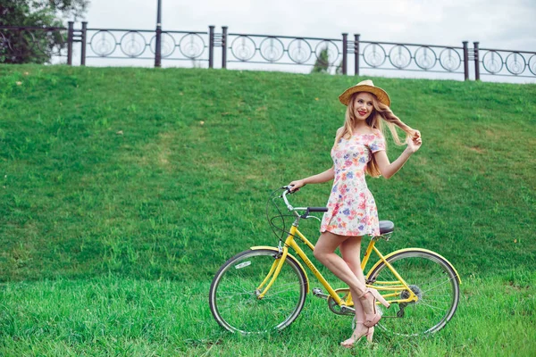 Retrato de comprimento total de uma bela mulher loira posando perto de uma bicicleta amarela em um parque de gramado verde — Fotografia de Stock