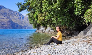 Vietnamlı genç bir kadın sırt çantasıyla kayanın üzerinde oturuyor ve gölün manzarasının tadını çıkarıyor.