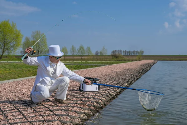 Exquisite fishing. Fisherman in white suite catch fish by spinning rod at lake — Stock Photo, Image