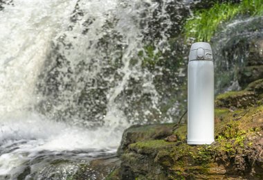 Beyaz termos çay ya da şelalenin yakınındaki taşın üzerinde su damlaları olan kahve. Macera yürüyüşü konsepti.