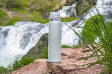 Beyaz termos çay ya da şelalenin yakınındaki taşın üzerinde su damlaları olan kahve. Macera yürüyüşü konsepti.