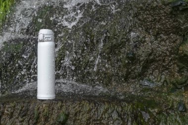 Beyaz termos çay ya da şelalenin yakınındaki taşın üzerinde su damlaları olan kahve. Macera yürüyüşü konsepti.