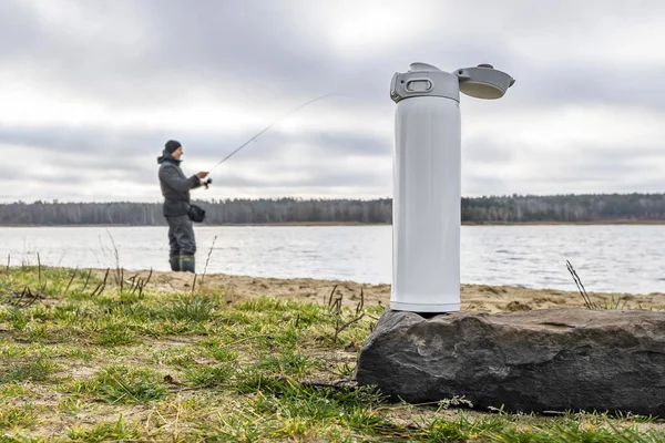 White thermos of tea or coffee on stone on lake background with fisherman silhouette. Fishing hiking concept. — Stock Photo, Image