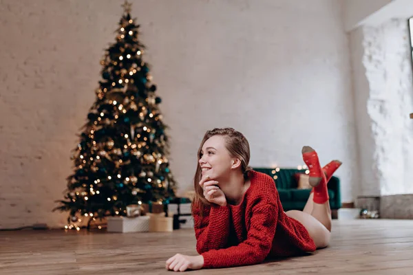 Młoda atrakcyjna kobieta leży na podłodze w czerwonym swetrze na tle noworocznego drzewa. — Zdjęcie stockowe