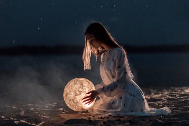 Gece kumsalında kumlu ve yıldızlı güzel bir genç kız Ay 'ı ellerinde tutuyor.