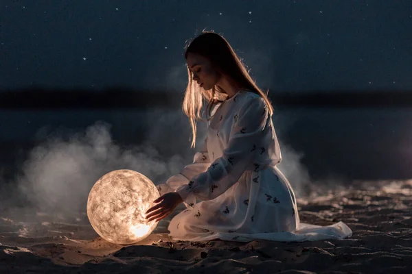 Piękna młoda dziewczyna na nocnej plaży z piaskiem i gwiazdami trzyma księżyc w rękach — Zdjęcie stockowe