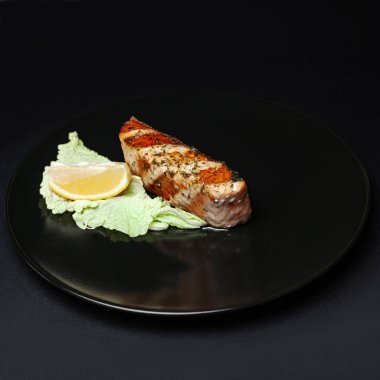 Siyah arka planda siyah tabakta salata ve limonla servis edilen somon bifteği..