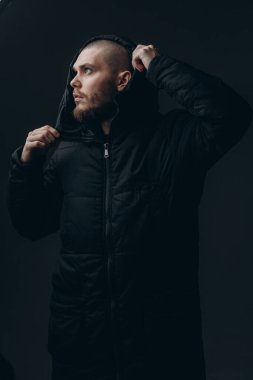 Gri arka planda duran, ceketli, ciddi bir gencin yakın plan portresi. Kel ve sakallı.