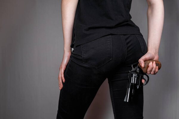 Armed girl in black jeans hides her pistols gun behind her back. Hidden threat. Killer woman.
