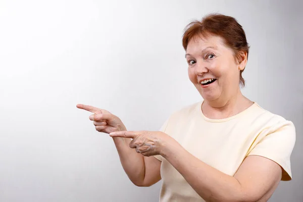 Beautiful senior woman shows a hand to the side on a white background in a light T-shirt, emotions — Stock Photo, Image