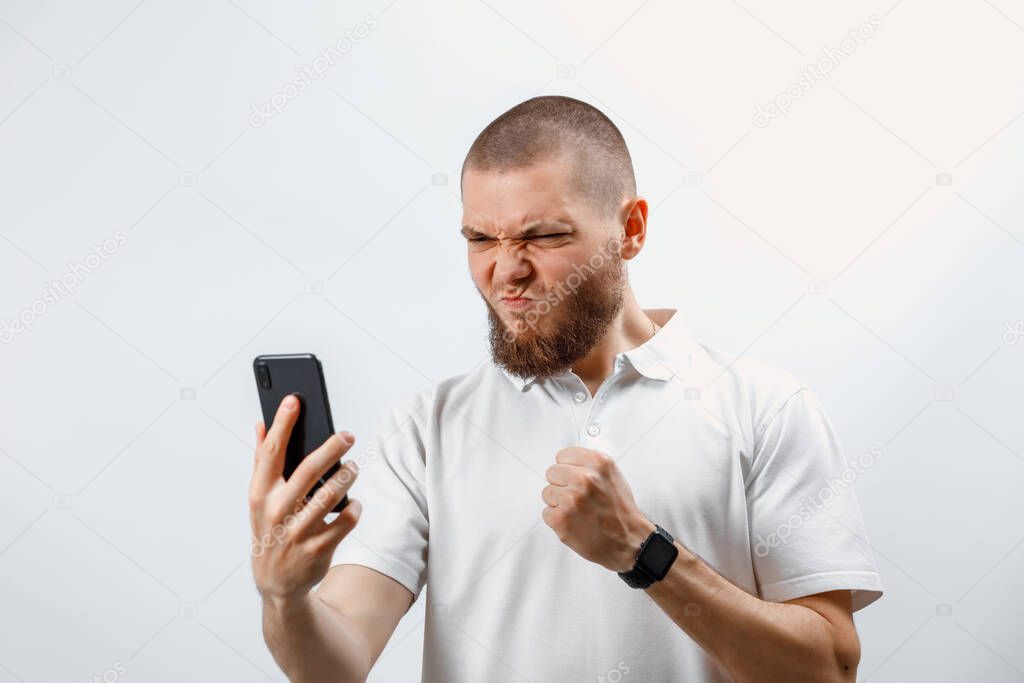 Portrait of an angry handsome bearded man in a white T-shirt holds a smartphone in his hand. isolated. emotions