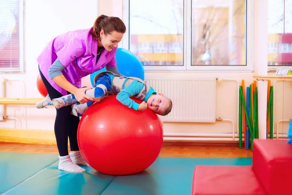 Lindo niño con discapacidad tiene terapia musculoesquelética haciendo ejercicios en cinturones de fijación del cuerpo en la bola en forma —  Fotos de Stock