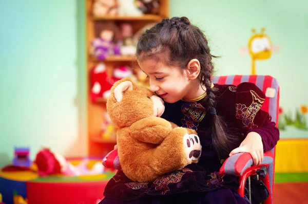 Милая маленькая девочка в инвалидной коляске обнимает плюшевого медведя в детском саду для детей с особыми потребностями — стоковое фото