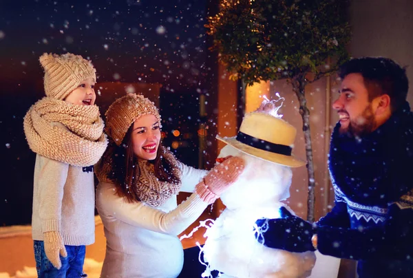 Feliz familia haciendo muñeco de nieve en la luz de la noche bajo nieve de invierno —  Fotos de Stock