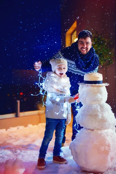 Счастливые отец и сын делают снеговика во дворе при свете фонарей — стоковое фото