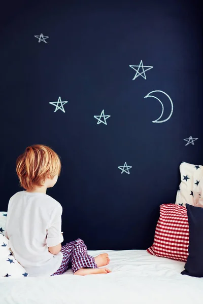 Cute kid in pajamas dreaming , while sitting in bed and looking on chalkboard wall — Stock Photo, Image
