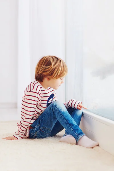 Niño guapo sentado en la alfombra cerca de la ventana en el día lluvioso —  Fotos de Stock