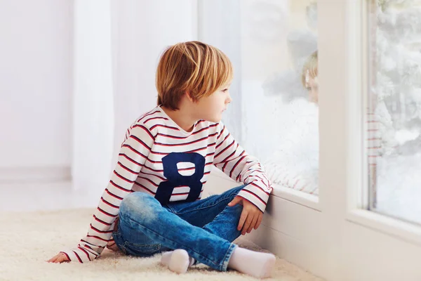 Stilig ung pojke sitter på mattan nära fönstret vid regnig dag — Stockfoto