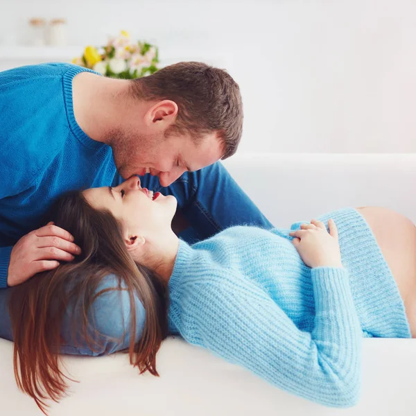 Счастливая беременная пара лежит дома на диване, целуется — стоковое фото