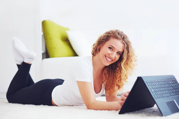 Feliz joven mujer que trabaja con el ordenador portátil, acostado en la alfombra en casa —  Fotos de Stock