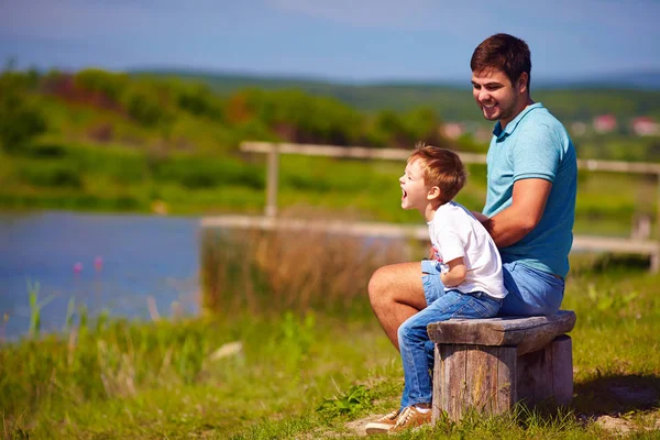 Ojciec i syn, dobra zabawa, siedząc na ławce nad jeziorem w słoneczny letni dzień — Zdjęcie stockowe
