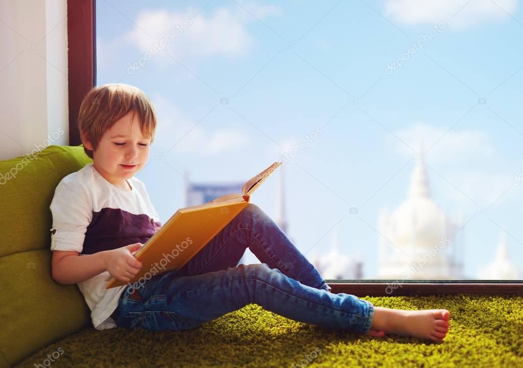 cute boy reading interesting book while sitting on carpet near the window at home