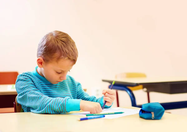 Menino bonito com necessidades especiais escrever cartas enquanto sentado na mesa na sala de aula — Fotografia de Stock
