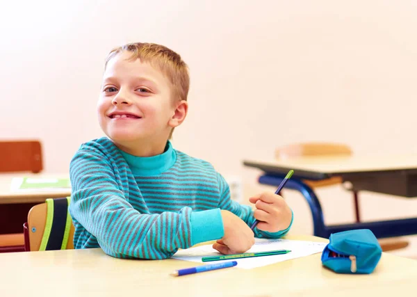 Mignon garçon avec des besoins spéciaux écrire des lettres tout en étant assis au bureau dans la salle de classe — Photo